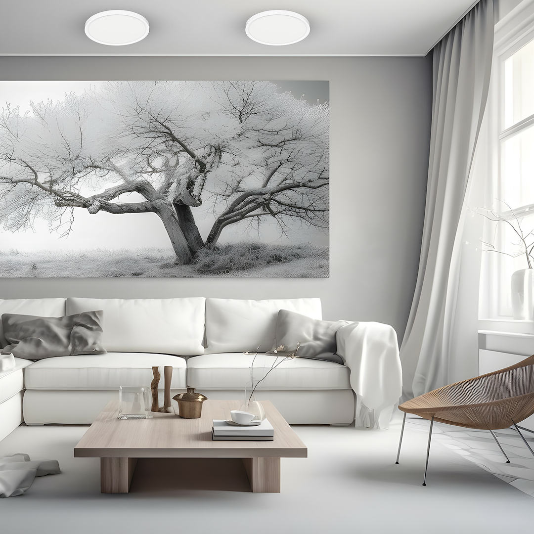 Plafon LED Maq D30 24W CCT w kolorze białym, okrągły, zamontowany na suficie w nowoczesnym, minimalistycznym wnętrzu. W tle widoczna szara ściana z obrazem drzew. Meble w jasnych kolorach, stół kawowy z akcesoriami.