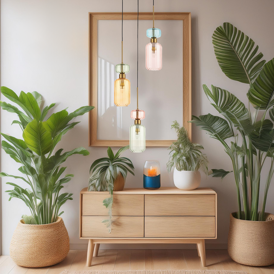 Lampa wisząca Molly 3xE14 w złotym kolorze z kolorowymi kloszami (zielony, różowy, pomarańczowy) zawieszona w poziomie nad drewnianą komodą, otoczona roślinami doniczkowymi, w jasnym wnętrzu z ramą obrazu w tle.