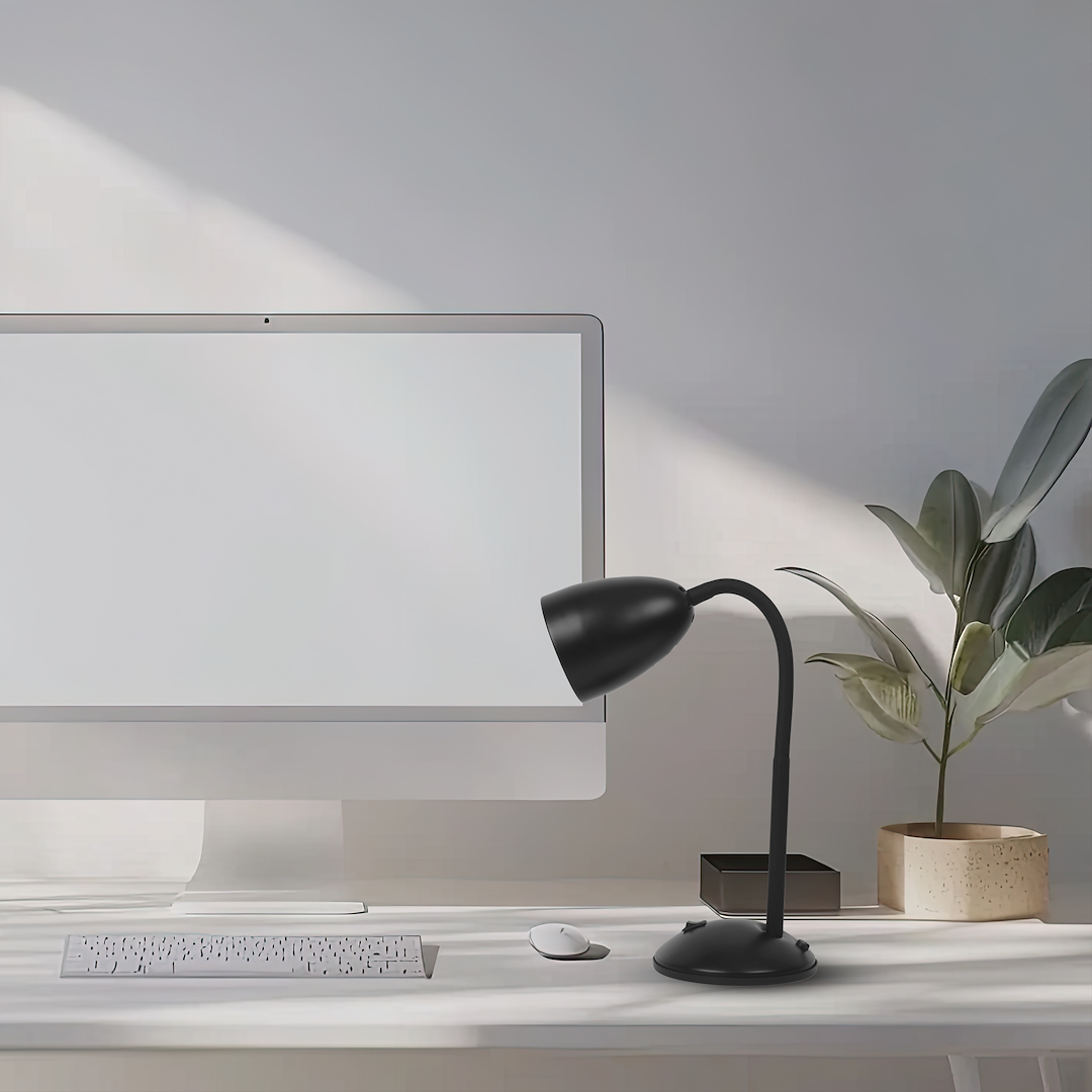 Czarna lampa biurkowa Logic z regulowanym kloszem, zamontowana na okrągłej podstawie. Ustawiona obok monitora i klawiatury na białym biurku, w tle widoczna roślina w doniczce.