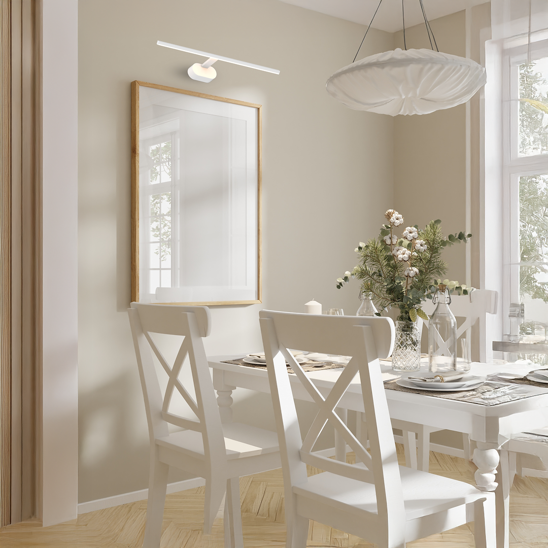 Kinkiet Prado L60 LED 11W 3000K w kolorze białym, zamontowany na ścianie nad białą ramą lustra. Oświetlenie w minimalistycznym stylu, idealne do aranżacji wnętrz. Widoczne elementy drewnianego stołu i krzeseł w jasnym kolorze.