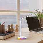 Lampa biurkowa LED Bruno ML1629 z białą obudową, z regulowanym ramieniem. Podstawa z kolorowym oświetleniem RGB. Widoczny laptop, drewniane figurki ptaków oraz notes z przyborami na biurku. Tło: okno z widokiem na morze.