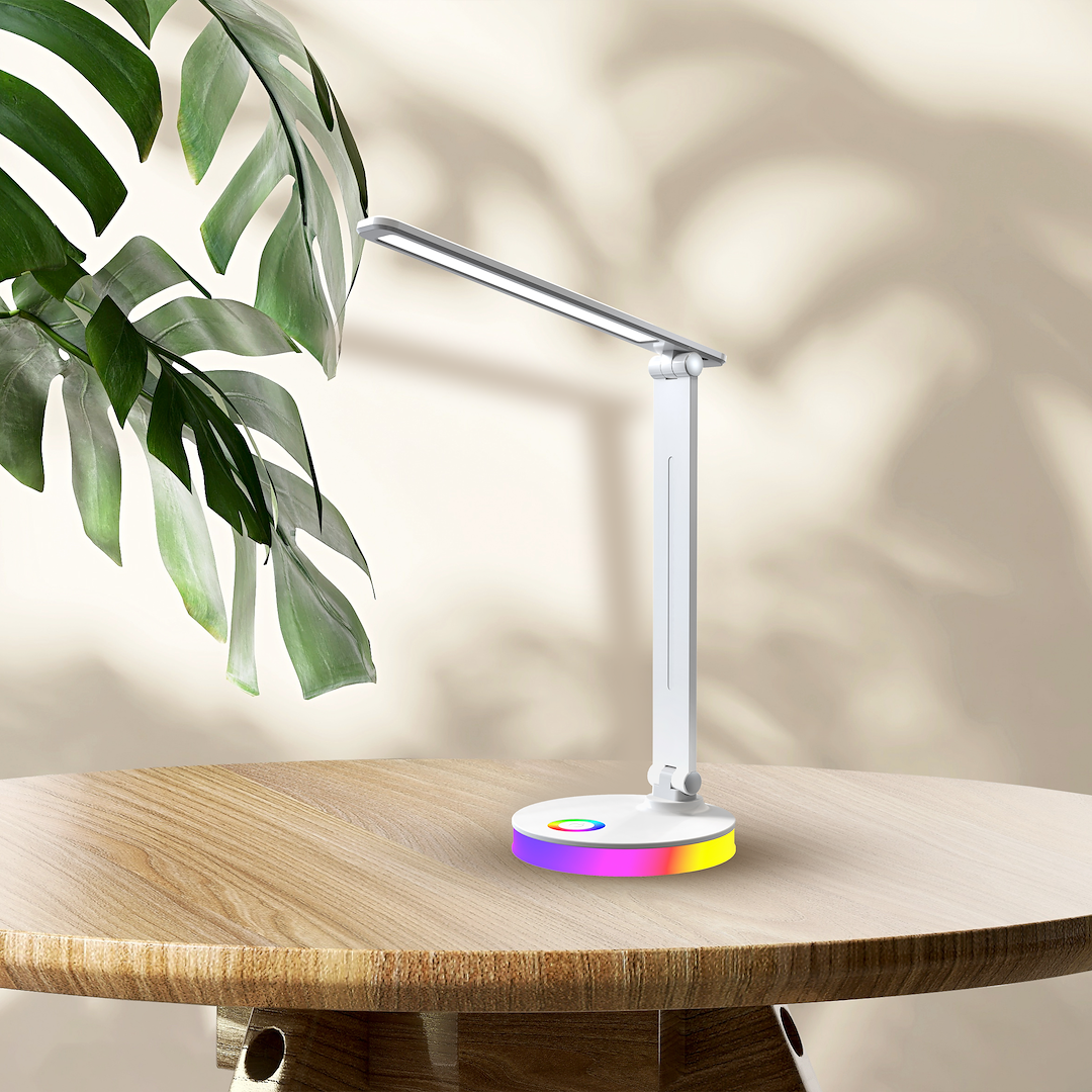 Bruno lampa biurkowa LED 5W CCT+RGB w białej obudowie, z regulowanym ramieniem. Podstawa z kolorowym podświetleniem. Ujęcie frontalne na drewnianym stole, w tle liście rośliny.