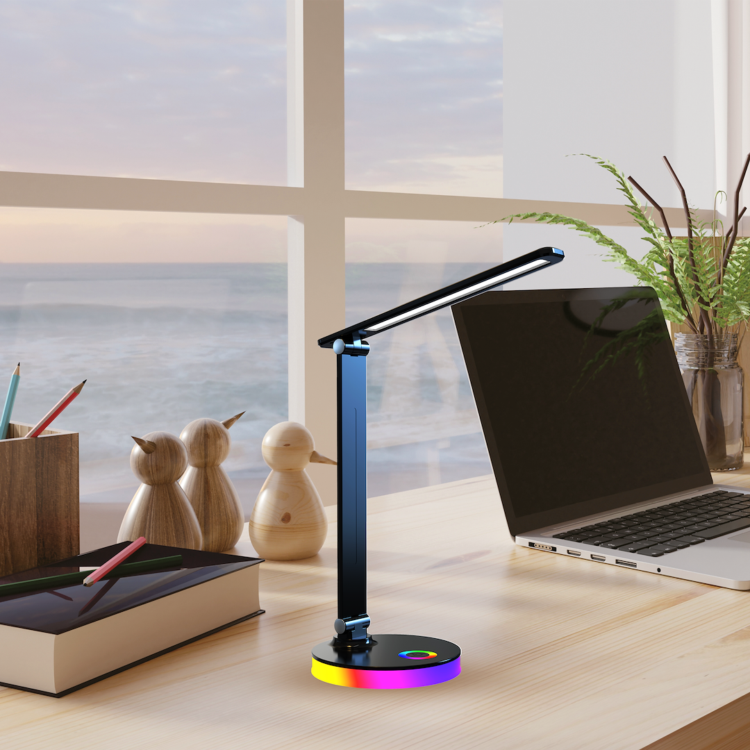 Czarna lampa biurkowa LED Bruno o mocy 5W, z funkcjami CCT i RGB. Posiada prostą, regulowaną konstrukcję z okrągłą podstawą w kolorach tęczy. Ustawiona na biurku obok laptopa i przyborów do pisania, w tle widoczne okno z widokiem na morze.