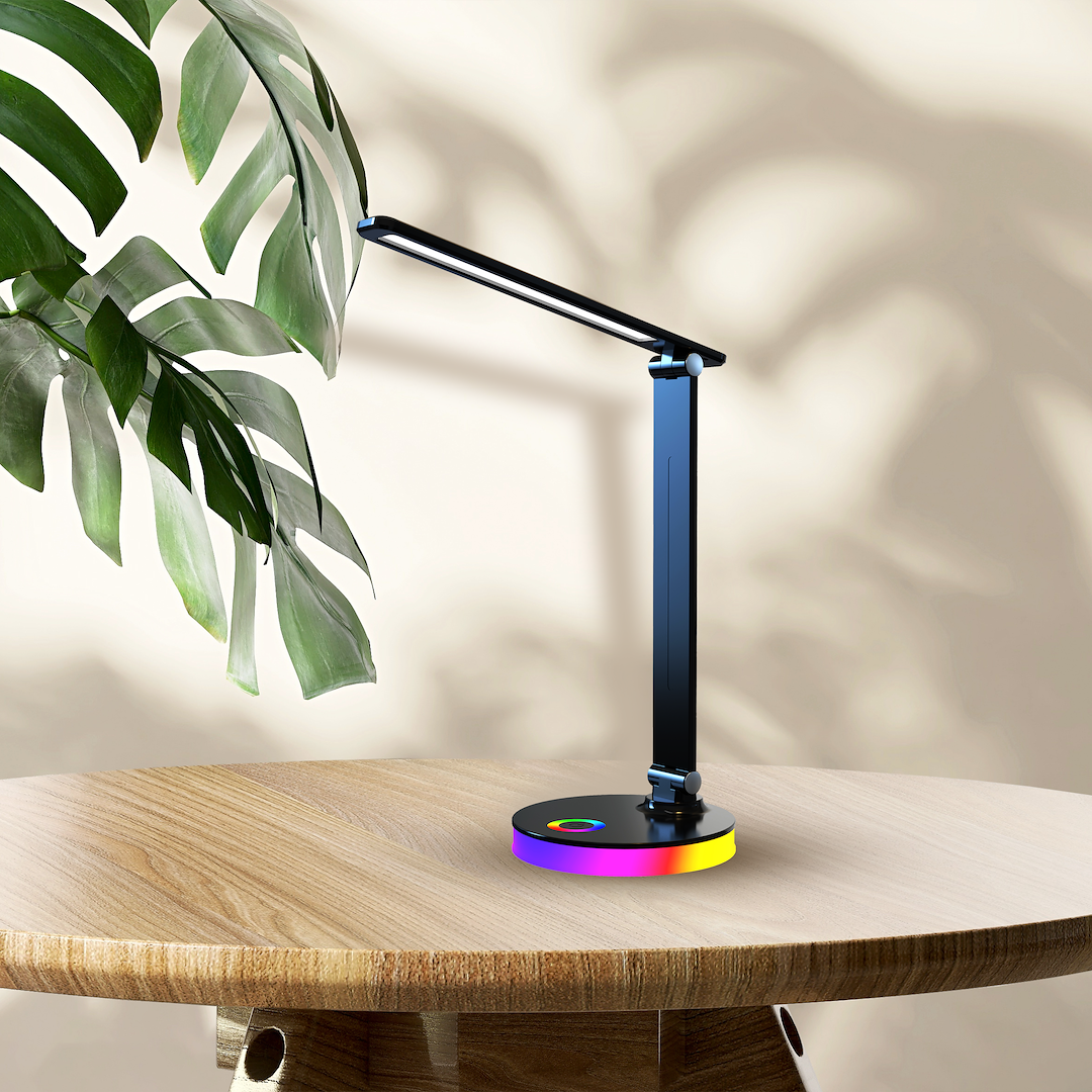 Czarna lampa biurkowa LED Bruno 5W CCT+RGB z regulowanym ramieniem, okrągłą podstawą z kolorowym podświetleniem. Ujęcie z boku na drewnianym stole, z tłem w odcieniach beżu i liściem rośliny w kadrze.