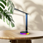 Czarna lampa biurkowa LED Bruno 5W CCT+RGB z regulowanym ramieniem, okrągłą podstawą z kolorowym podświetleniem. Ujęcie z boku na drewnianym stole, z tłem w odcieniach beżu i liściem rośliny w kadrze.