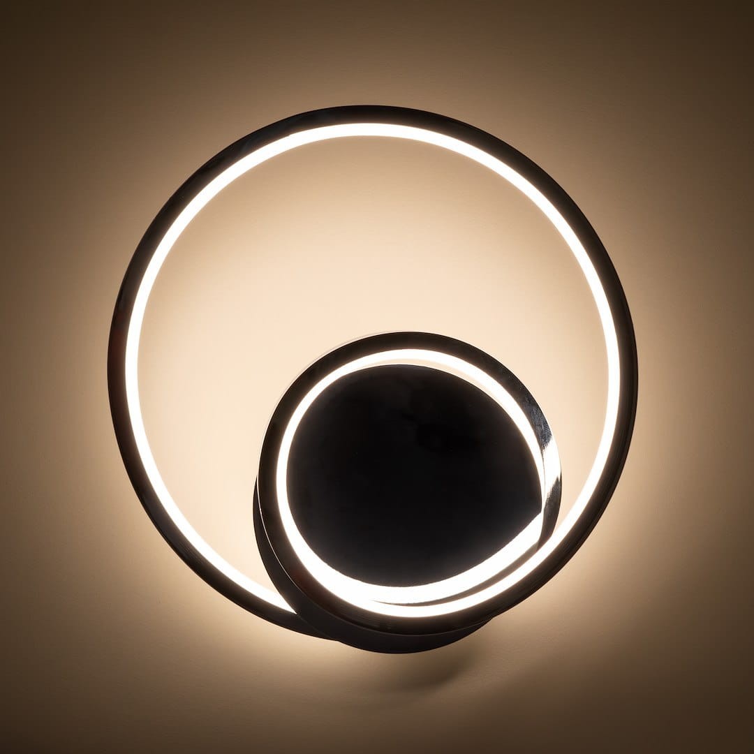 Kinkiet LED Lucero 13W 4000K w kolorze chromu, złożony z dwóch okrągłych elementów, jeden większy zewnętrzny i mniejszy wewnętrzny, emitujący ciepłe światło. Montowany na ścianie, z minimalistycznym designem, na jednolitym tle.