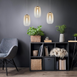 Lampa wisząca LED Vaso 3 z trzema kloszami w złotym i bursztynowym odcieniu, zawieszona nad czarną półką. Klosze mają owalny kształt, a całość otoczona zielonymi roślinami oraz nowoczesnym meblem.