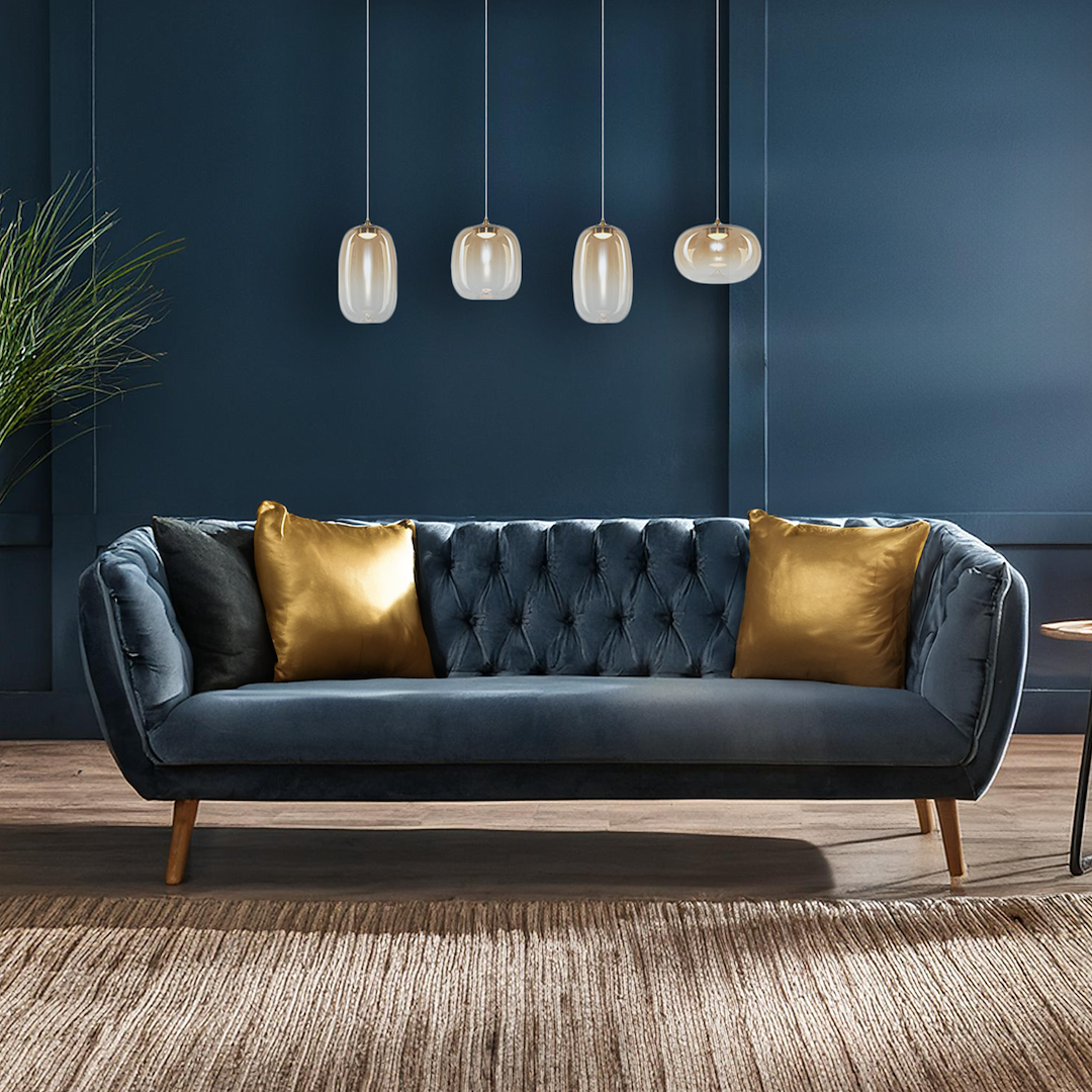 Lampa wisząca LED Vaso 15W 3000K w złotym i bursztynowym kolorze, z eleganckim, zaokrąglonym kształtem. Zawieszona w grupie nad niebieską sofą z poduszkami w kolorze złotym i czarnym, w nowoczesnym wnętrzu.