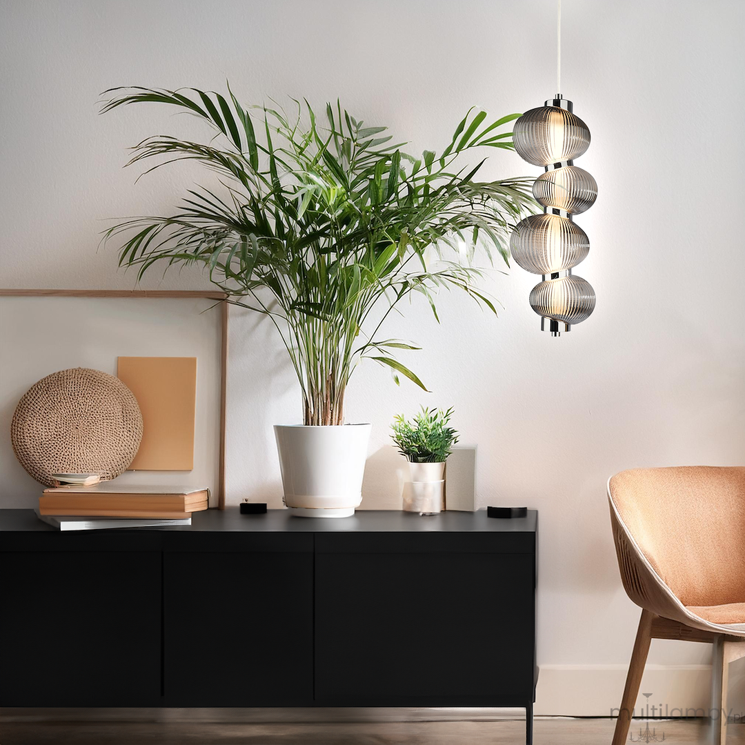 Lampa wisząca LED Bolla 17W 3000K w kolorze chromu z dymionymi kloszami, zawieszona nad ciemnym stolikiem w nowoczesnym wnętrzu. Otoczona roślinami w doniczkach, z elementami dekoracyjnymi w tle.