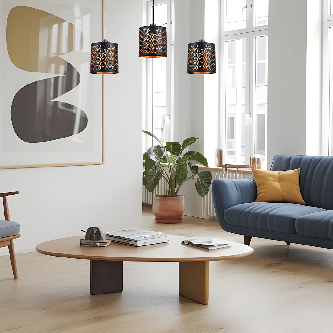 Lampa wisząca Morgan z trzema źródłami światła E27 w czarnej obudowie z ażurowym wzorem, zawieszona w salonie nad okrągłym stołem. W tle widoczne okna oraz roślina w doniczce, a także meble w stonowanych kolorach.