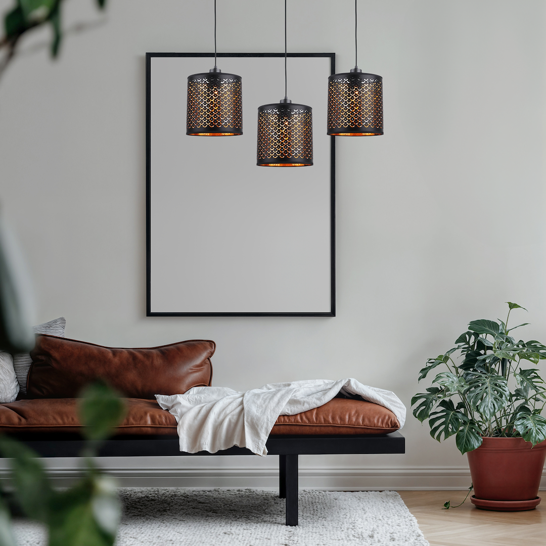Lampa wisząca Morgan w kolorze czarnym z trzema kloszami E27, o ażurowym wzorze, zawieszona na długich kablach. W tle widoczna kanapa w odcieniu brązu oraz roślina doniczkowa.
