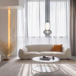 Lampa wisząca LED Arame 15W 3000K w kolorze czarnym, zawieszona nad białą sofą. Okrągła podstawa z siatki metalowej oraz szklany klosz w kształcie butelki. W tle jasne zasłony i stół z przezroczystym blatem.