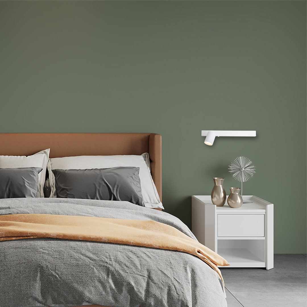 Kinkiet Magnet 1xGU10 w białym kolorze, zamontowany na zielonej ścianie obok łóżka. Oświetlenie skierowane w dół, z lampą LED 5W CCT. W tle widoczne poduszki, narzuta w odcieniach szarości oraz nocna szafka z dekoracjami.