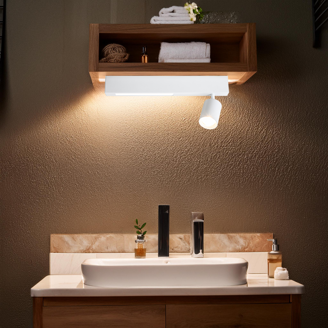 Lampa sufitowa Magnet 1xGU10 + LED 5W CCT w kolorze białym, zamontowana na ścianie nad umywalką. Oświetlenie skierowane w dół, z minimalistyczną, cylindryczną oprawą. W tle drewniana półka z ręcznikami i kosmetykami, w stonowanej aranżacji.