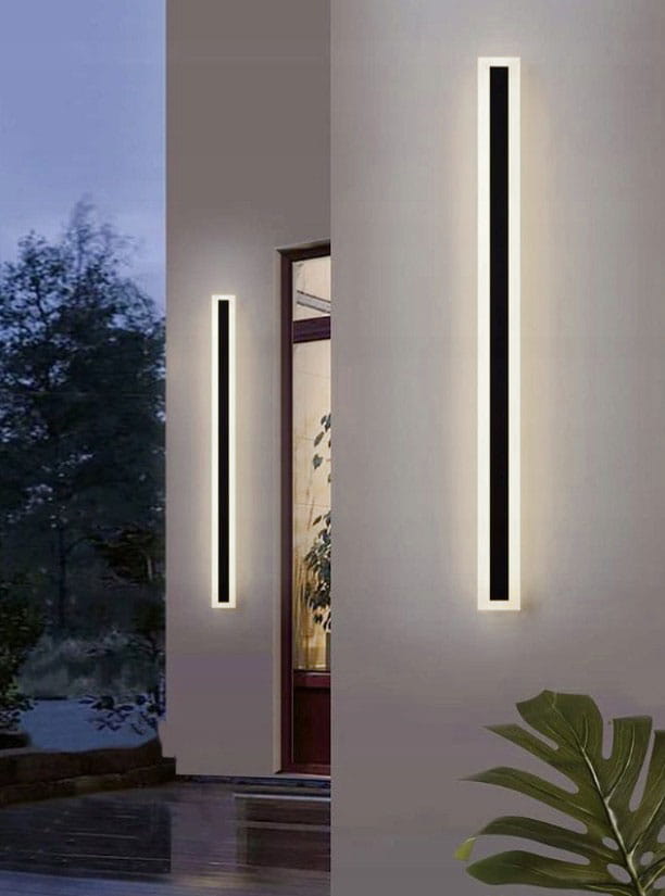 Kinkiet elewacyjny LED Durango 100, czarny, o prostokątnym kształcie. Dwa długie paski LED emitują światło o temperaturze barwowej 4500K. Zainstalowany na zewnętrznej ścianie, obok drzwi, na tle roślinności.
