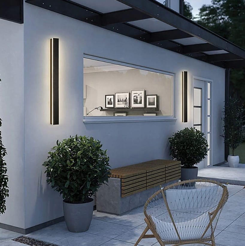 Czarny kinkiet elewacyjny LED Lara 150 o mocy 45W i temperaturze barwowej 4000K, montowany na ścianie, z prostokątnym kształtem. W tle widoczne rośliny w donicach oraz drewniana ławka, w aranżacji nowoczesnego zewnętrznego wejścia.