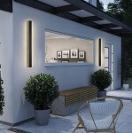 Czarny kinkiet elewacyjny LED Lara 150 o mocy 45W i temperaturze barwowej 4000K, montowany na ścianie, z prostokątnym kształtem. W tle widoczne rośliny w donicach oraz drewniana ławka, w aranżacji nowoczesnego zewnętrznego wejścia.