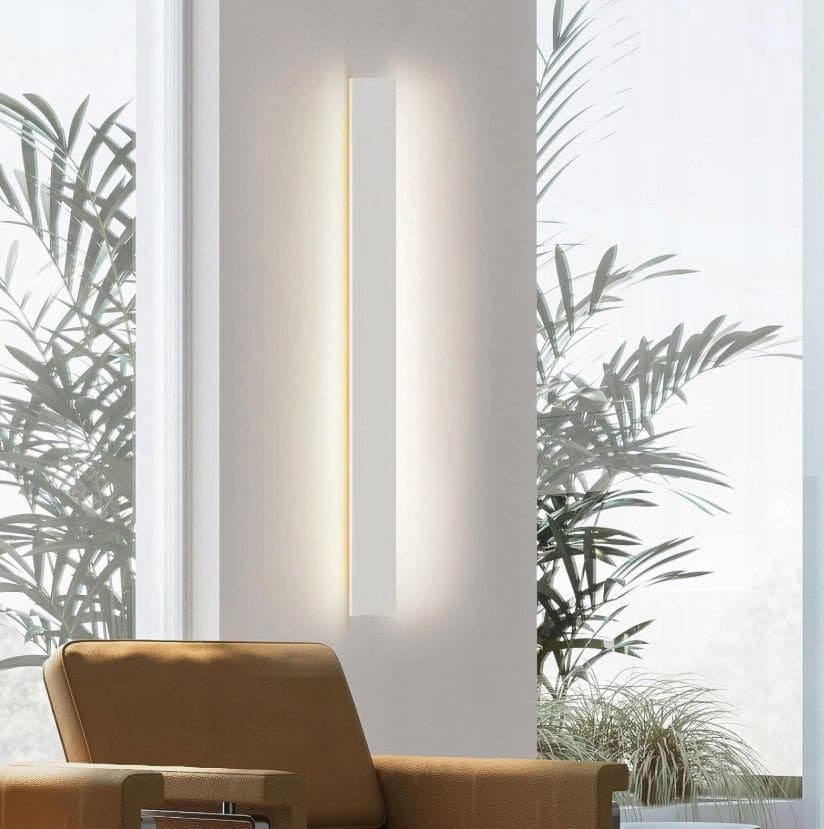 Kinkiet elewacyjny LED Lara 100 o mocy 30W i barwie 4000K w białej obudowie. Montowany pionowo na ścianie, emituje ciepłe światło. W tle widoczna roślina doniczkowa oraz fragment nowoczesnego wnętrza.