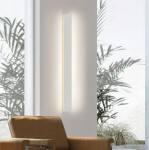 Kinkiet elewacyjny LED Lara 100 o mocy 30W i barwie 4000K w białej obudowie. Montowany pionowo na ścianie, emituje ciepłe światło. W tle widoczna roślina doniczkowa oraz fragment nowoczesnego wnętrza.