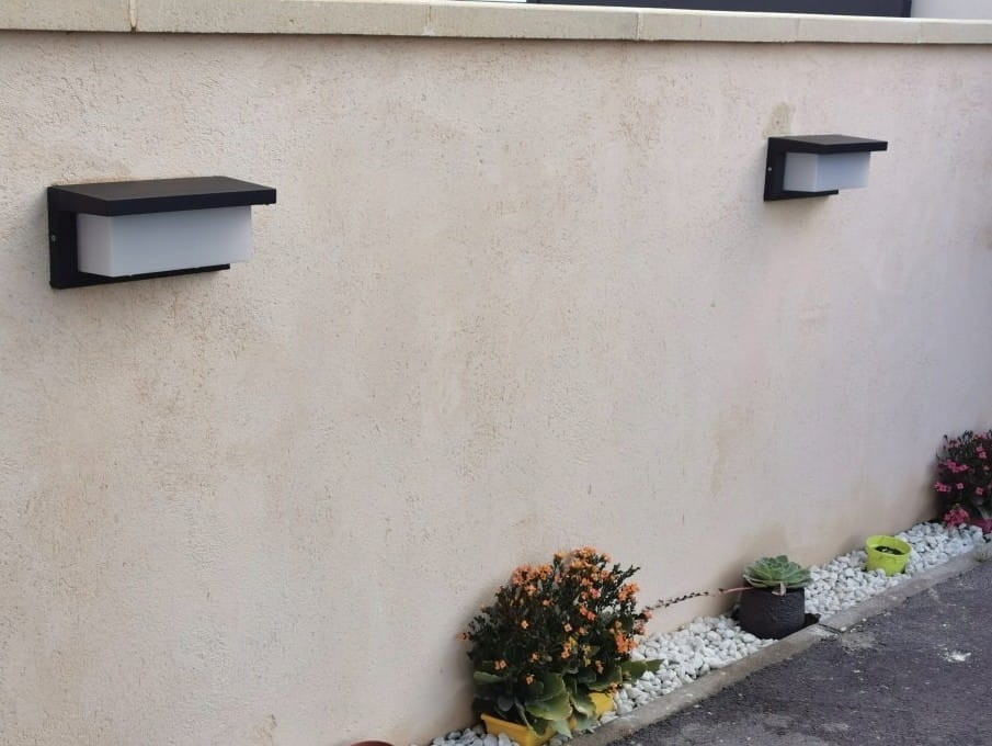 Lagos kinkiet elewacyjny w kolorze czarnym, montowany na ścianie, z prostokątnym kloszem z białego szkła. Widoczny w aranżacji z roślinami doniczkowymi oraz żwirem, tworzy nowoczesny akcent oświetleniowy.