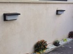 Lagos kinkiet elewacyjny w kolorze czarnym, montowany na ścianie, z prostokątnym kloszem z białego szkła. Widoczny w aranżacji z roślinami doniczkowymi oraz żwirem, tworzy nowoczesny akcent oświetleniowy.