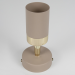 Kinkiet Joker w kolorze kaszmiru ze złotym wykończeniem. Cylindryczny klosz na podstawie o okrągłym kształcie, z widocznym mocowaniem. Lampa przeznaczona na żarówkę GU10, układ frontalny. Tło białe, minimalistyczne.