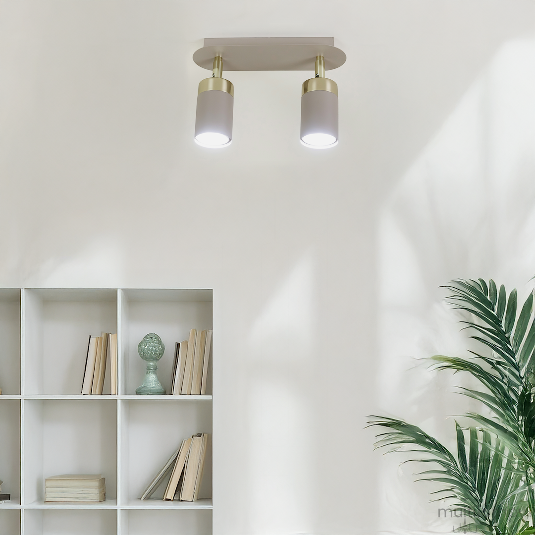 Lampa sufitowa Joker w kolorze kaszmiru i złota z dwoma reflektorami GU10, zamontowana na prostokątnej podstawie. Widoczna w jasnym wnętrzu z regałem na książki i rośliną w tle. Reflektory emitują światło skierowane w dół.