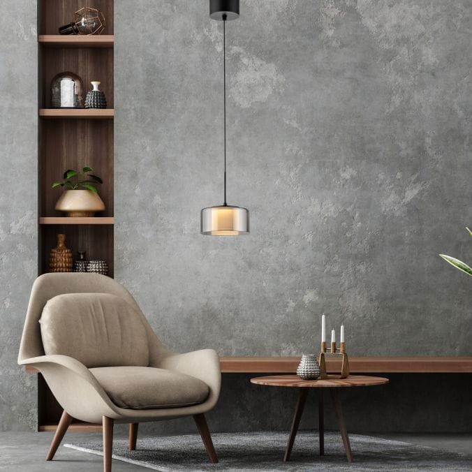 Lampa wisząca LED Rika 6W w kolorze czarnym z dymionym kloszem, zawieszona na cienkim przewodzie. Produkt umieszczony w nowoczesnym wnętrzu z beżowym fotelem i drewnianym stolikiem, na tle szarej ściany z półkami.