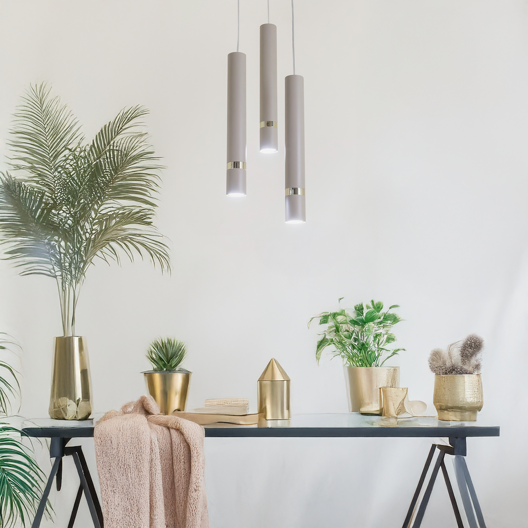Lampa wisząca Joker 3xGU10 w kolorze kaszmiru i złota, zawieszona na cienkich linkach. Składa się z trzech cylindrycznych kloszy, umieszczonych w różnych wysokościach. Otoczenie to stół z roślinami w złotych donicach i dekoracjami, na jasnym tle.