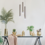 Lampa wisząca Joker 3xGU10 w kolorze kaszmiru i złota, zawieszona na cienkich linkach. Składa się z trzech cylindrycznych kloszy, umieszczonych w różnych wysokościach. Otoczenie to stół z roślinami w złotych donicach i dekoracjami, na jasnym tle.