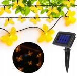 Girlanda solarna o długości 580 cm z 20 LED w kształcie żółtych motylków. Motylki umieszczone na czarnym przewodzie, z solarnym panelem w kształcie prostokąta. Elementy dekoracyjne idealne do ogrodu, na tle zielonych liści.