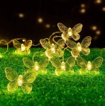 Girlanda ogrodowa o długości 580 cm z 20 LED w kształcie motylków. Motylki w kolorze przezroczystym z delikatnym wzorem, umieszczone na cienkim przewodzie, leżą na zielonej trawie, emitując ciepłe światło. Tło ciemne, co podkreśla ich efekt świetlny.