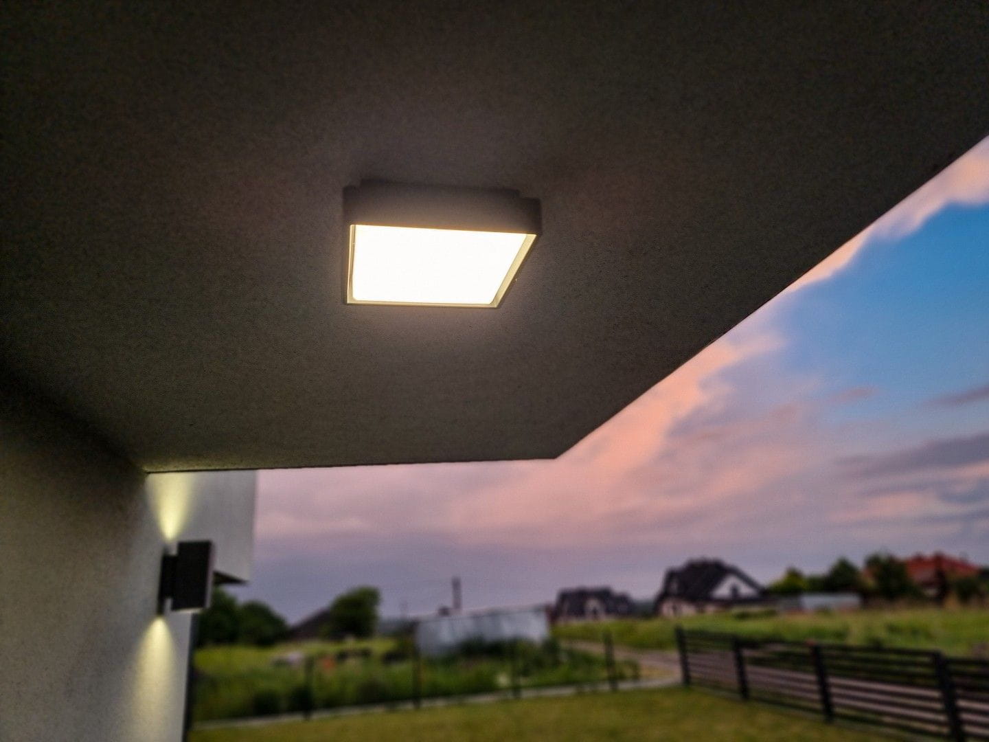 Plafon elewacyjny Beni D20x20 LED 18W CCT w kolorze ciemnego popielu, zamontowany pod sufitem. Widoczny prostokątny kształt oraz emitowane światło na tle zachmurzonego nieba i zielonej trawy.