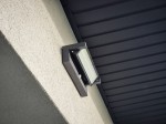 Kinkiet elewacyjny kierunkowy w ciemnym popiele z czujnikiem ruchu. Ujęcie z boku, zamontowany na ścianie, z widoczną matową szybą LED. Oświetlenie o mocy 27W, barwa 4000K, skierowane w dół. Tło jasne, gładkie.