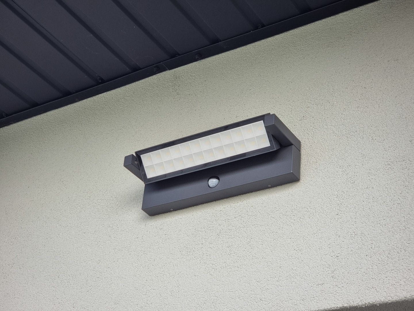 Kinkiet elewacyjny kierunkowy LED 27W, ciemny popiel, z czujnikiem ruchu. Umieszczony na ścianie, o prostokątnym kształcie z białym kloszem, emitującym światło o temperaturze 4000K. Widoczny element montażowy i tekstura powierzchni ściany.