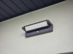 Kinkiet elewacyjny kierunkowy LED 27W, ciemny popiel, z czujnikiem ruchu. Umieszczony na ścianie, o prostokątnym kształcie z białym kloszem, emitującym światło o temperaturze 4000K. Widoczny element montażowy i tekstura powierzchni ściany.