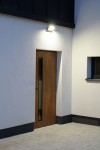 Kinkiet elewacyjny kierunkowy LED 21W 4000K w ciemnym popiele, zamontowany na ścianie obok drewnianych drzwi. Oświetlenie skierowane w dół, z czujnikiem ruchu, w otoczeniu jasnej, gładkiej elewacji budynku.