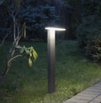 Lampa stojąca ogrodowa LED Fler H78 w kolorze antracytowym, o wysokości 78 cm i mocy 10W, emituje światło o temperaturze 4000K. Ustawiona w ogrodzie, obok roślinności, z prostokątnym kloszem na górze, na tle ciemnego otoczenia.