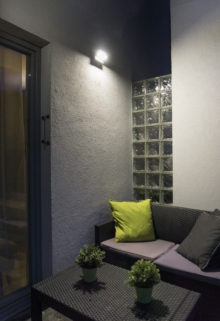 Kinkiet elewacyjny Rota LED RT-K100 w kolorze srebrnym, zamontowany na ścianie, kierunkowy, o prostokątnym kształcie. Oświetla strefę wypoczynkową z sofą i poduszkami w zielonym i szarym kolorze, otoczony roślinami w doniczkach.