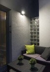 Kinkiet elewacyjny Rota LED RT-K100 w kolorze srebrnym, zamontowany na ścianie, kierunkowy, o prostokątnym kształcie. Oświetla strefę wypoczynkową z sofą i poduszkami w zielonym i szarym kolorze, otoczony roślinami w doniczkach.