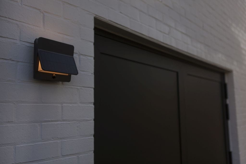 Kinkiet solarny LED Aleta 12,6 W w kolorze antracytowym, zamontowany na białej ścianie. Posiada panel słoneczny na górze oraz źródło światła emitujące ciepłą barwę 3000K. Widoczny zbliżony kąt ujęcia.