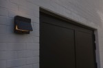 Kinkiet solarny LED Aleta 12,6 W w kolorze antracytowym, zamontowany na białej ścianie. Posiada panel słoneczny na górze oraz źródło światła emitujące ciepłą barwę 3000K. Widoczny zbliżony kąt ujęcia.