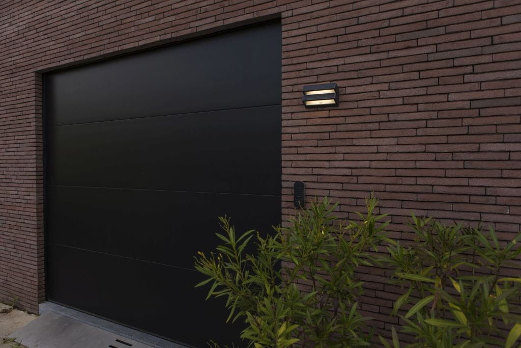 Kinkiet solarny LED Barlo o mocy 8,2 W w kolorze antracytowym. Zamontowany na elewacji budynku, z dwiema poziomymi żarówkami na tle ciemnej bramy i ceglanej ściany. Roślinność w dolnej części kadru.