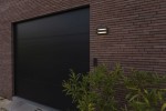 Kinkiet solarny LED Barlo o mocy 8,2 W w kolorze antracytowym. Zamontowany na elewacji budynku, z dwiema poziomymi żarówkami na tle ciemnej bramy i ceglanej ściany. Roślinność w dolnej części kadru.