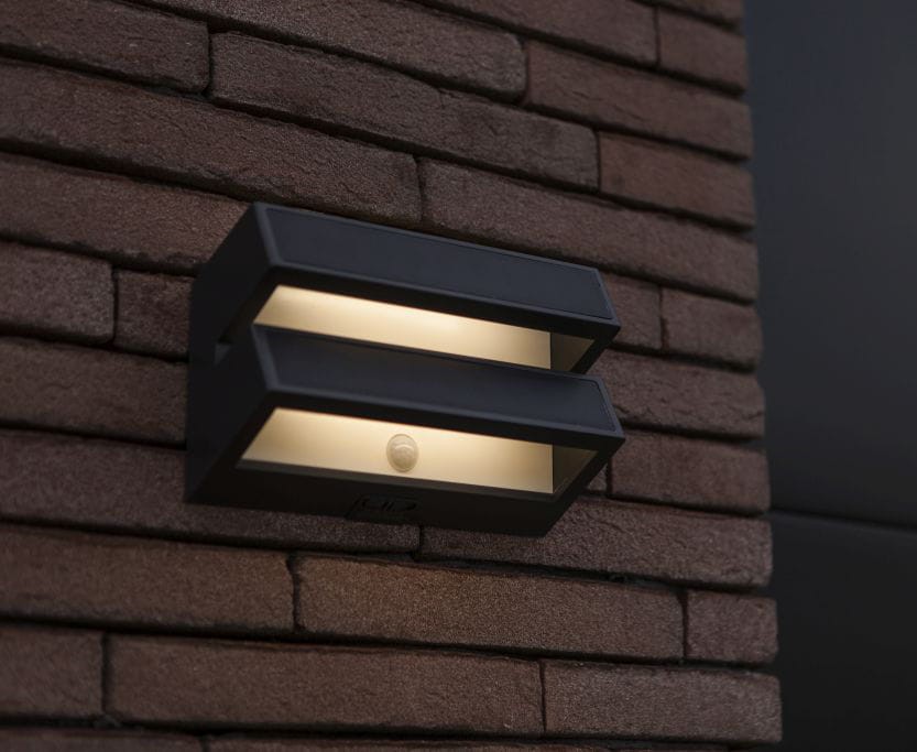 Kinkiet solarny LED Barlo w kolorze antracytowym, z dwiema poziomymi szczelinami emitującymi światło, zamontowany na ścianie z cegły. Widoczny czujnik ruchu na dolnej części. Oświetlenie o temperaturze barwowej 4000K, nowoczesny design.