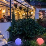 Kula D30, solarna lampa ogrodowa LED RGB, o kulistym kształcie, umieszczona w ogrodzie. Widoczne są dwa egzemplarze w kolorach różowym i niebieskim, otoczone zielenią roślinności. Tło to oświetlony taras i dom.