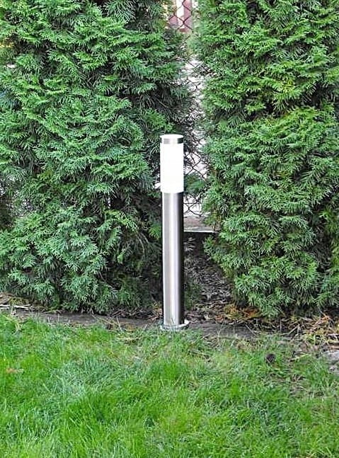 Okrągła lampa stojąca ogrodowa Manila H65 w kolorze srebrnym, z białym kloszem, umieszczona między dwoma zielonymi krzewami. Lampa ma prosty, cylindryczny kształt, stojąca na trawniku.