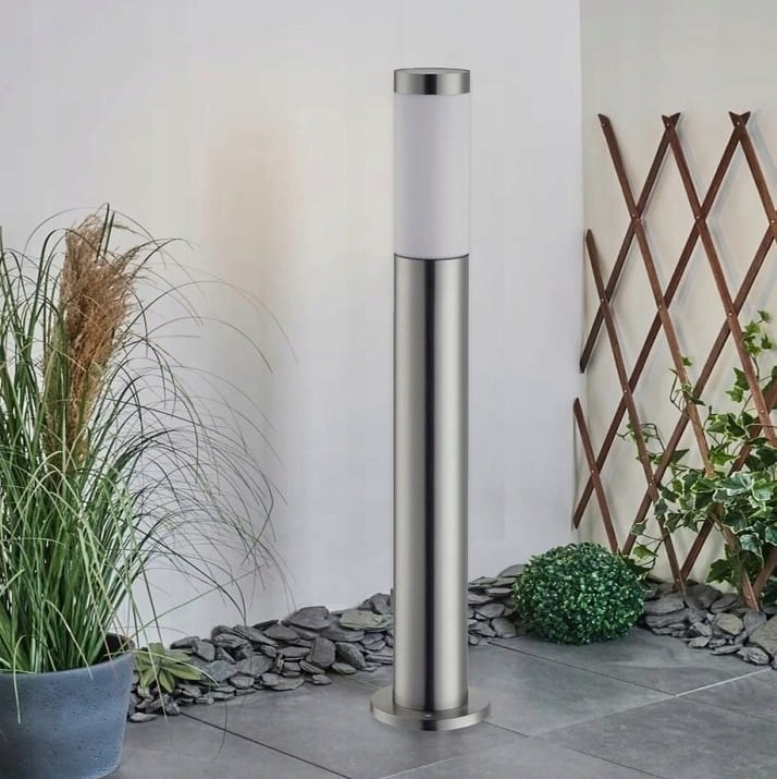 Lampa stojąca Manila H65 w formie okrągłego słupka, wykonana ze srebrnego metalu, z białym kloszem na górze. Ustawiona na płaskiej powierzchni, otoczona roślinnością, w nowoczesnym ogrodzie.
