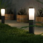 Czarna lampa stojąca Manila H45, okrągła, z białym kloszem, umieszczona na trawie. W tle widoczne drewniane donice z roślinami. Lampa emitująca światło, z prostym, nowoczesnym designem, ustawiona w ogrodzie.