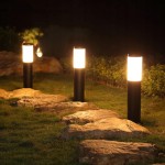 Czarna lampa stojąca słupek Manila H45, okrągła, umieszczona na trawie w ogrodzie, z emitującym światło kloszem. Ujęcie ukazuje trzy lampy, każda na stabilnym podłożu, w otoczeniu kamieni i niskiej roślinności.